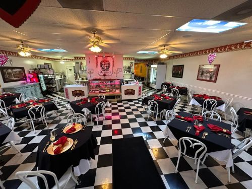 A cozy restaurant interior with black and white checkered floors, tables set with red napkins, and a pink dessert display.