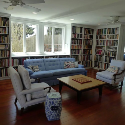 Cozy living room with a blue sofa, armchairs, wooden coffee table, and bookshelves filled with books. Bright natural light.