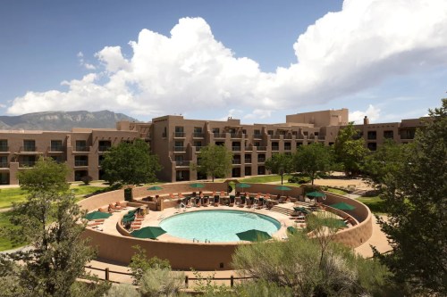 A circular pool surrounded by lounge chairs and green umbrellas, with a hotel and mountains in the background under a blue sky.