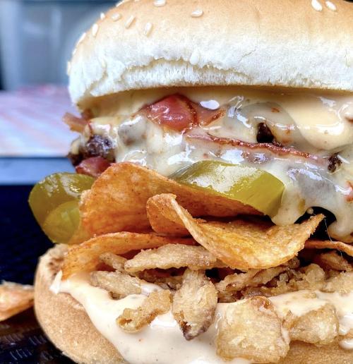Close-up of a sesame seed bun burger layered with chips, pickles, tomatoes, and creamy sauce.