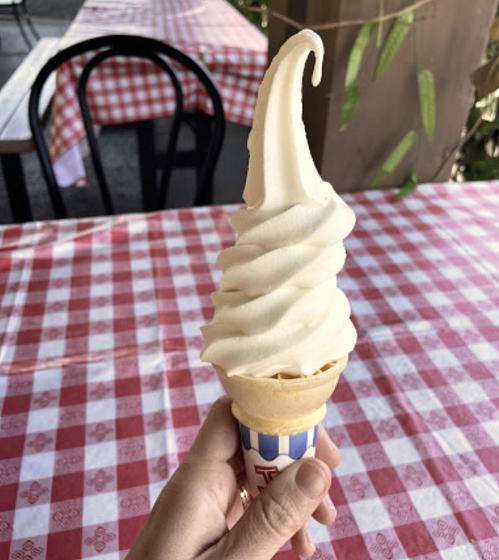 A hand holding a soft-serve ice cream cone with a swirl of creamy vanilla on a checkered tablecloth.