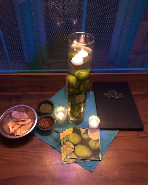 A table setting with a tall vase of water and limes, candles, a menu, and bowls of chips and sauces.