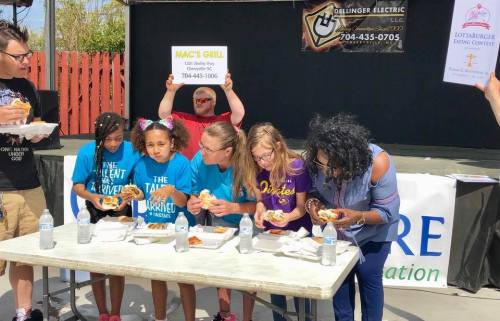 A group of children and adults participate in a food contest at an outdoor event, enjoying burgers and drinks.