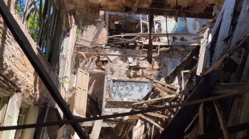Interior of a dilapidated building with exposed walls, broken beams, and debris scattered throughout.