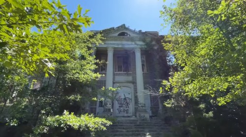 Abandoned building with columns, overgrown with trees and vines, under a clear blue sky.