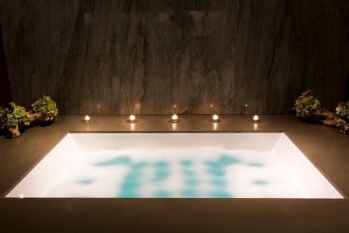 A modern bathtub filled with water, surrounded by candles and decorative plants against a textured wall.