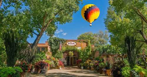 A colorful hot air balloon floats above a charming building surrounded by lush greenery and vibrant flower pots.