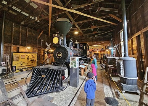 A vintage steam locomotive displayed in a museum, with visitors exploring the exhibit in a rustic wooden building.