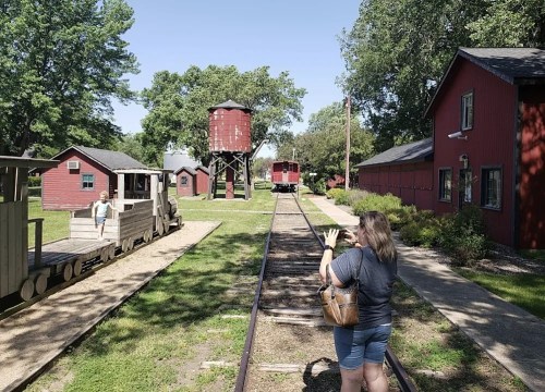 A woman stands on a train track, looking at a vintage train station and red buildings under a clear blue sky.