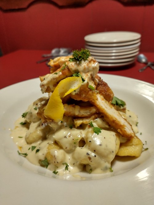 A plate of creamy pasta topped with fried chicken, lemon, and herbs, served on a red tablecloth with stacked plates in the background.