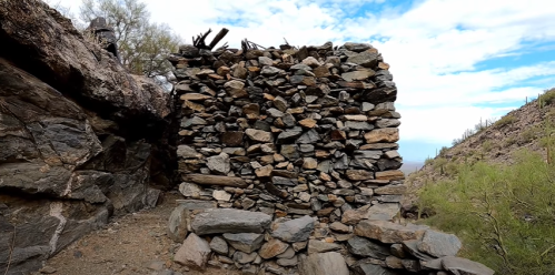 A stone structure made of stacked rocks, surrounded by desert landscape and mountains under a cloudy sky.