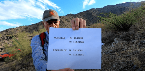 A person holds a paper with GPS coordinates for a trailhead and rock house against a mountainous background.
