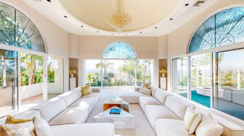 Bright, spacious living room with large windows, modern furniture, and a view of greenery outside. Elegant chandelier above.