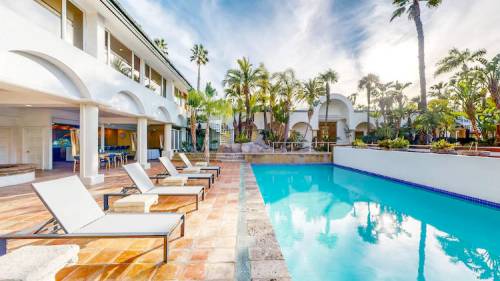 A serene pool area with lounge chairs, surrounded by palm trees and a stylish building under a bright sky.