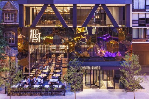 Modern hotel exterior featuring a reflective facade, outdoor seating, and vibrant lighting in an urban setting.