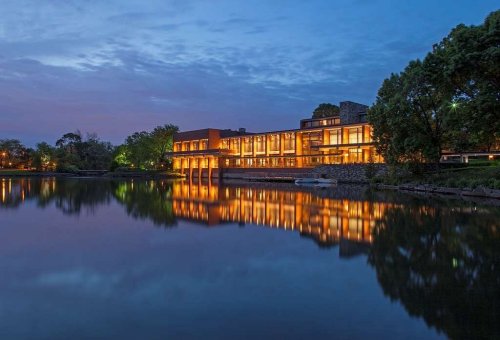 A modern building by a lake at dusk, with warm lights reflecting on the water's surface and trees in the background.