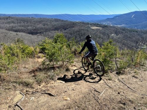 A mountain biker rides along a trail with a scenic view of hills and valleys under a clear blue sky.