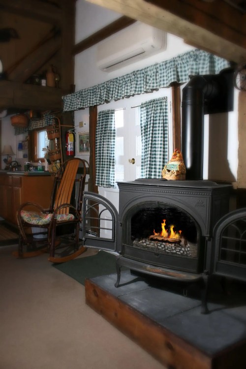 Cozy interior of a cabin featuring a rocking chair, a wood stove with flames, and a rustic kitchen area.