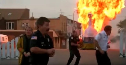 A group of police officers runs away from a large explosion in a town setting, with flames rising in the background.