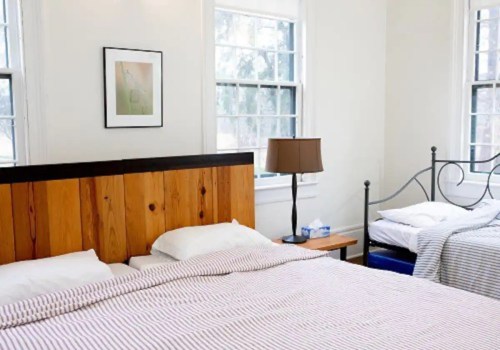 Cozy bedroom with a wooden headboard, two beds, a lamp, and large windows letting in natural light.