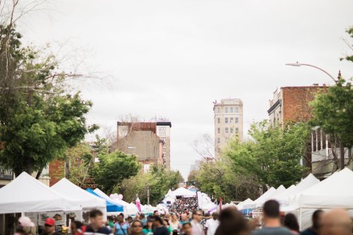 A bustling street fair with tents lining the road and crowds of people, set against a cloudy sky and city buildings.