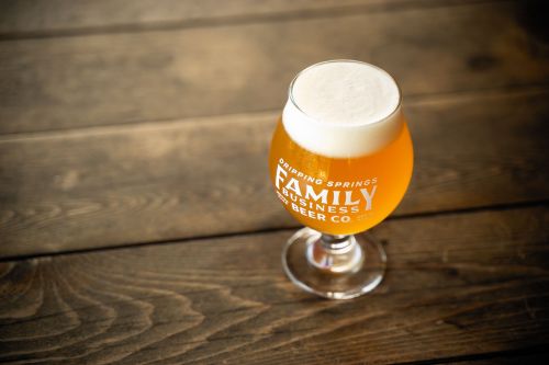 A glass of beer with a frothy head, resting on a wooden table, featuring the logo of Family Business Beer Co.