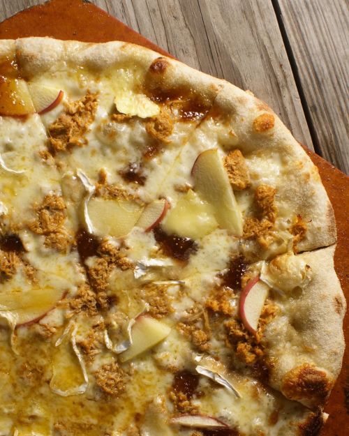 A close-up of a pizza topped with cheese, apple slices, and a drizzle of sauce on a wooden surface.