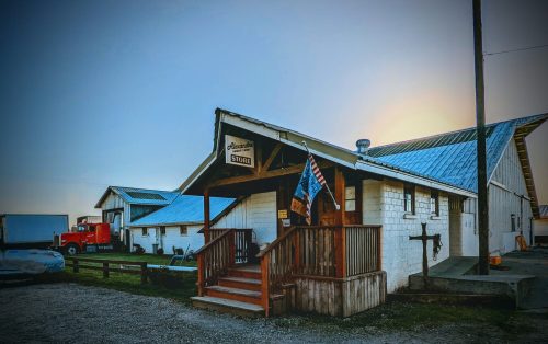 A rustic store with a wooden porch, surrounded by farm buildings and a red truck parked nearby under a clear sky.