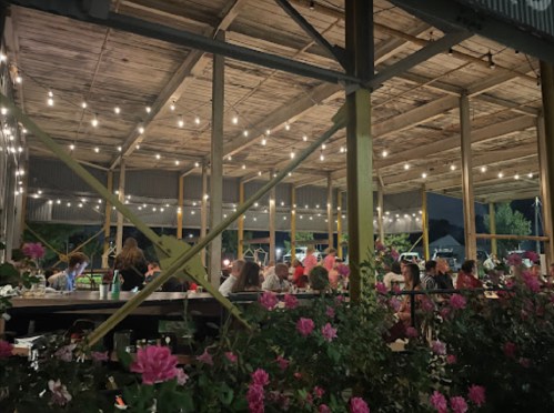 A lively outdoor dining area at night, adorned with string lights and surrounded by blooming flowers.