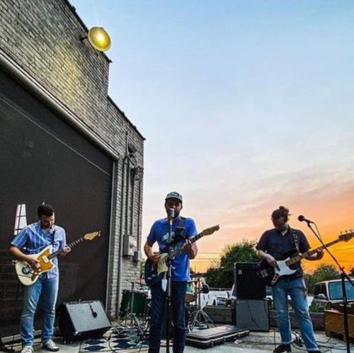 Three musicians perform on an outdoor stage at sunset, playing guitars with amplifiers and a backdrop of a brick wall.