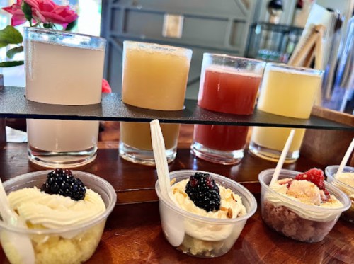 A wooden tray displays colorful drinks in glasses and small cups of desserts topped with berries.