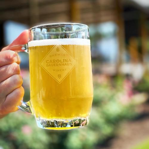 A hand holding a clear glass mug filled with light beer, featuring a logo for Carolina Bauernhaus Winery.