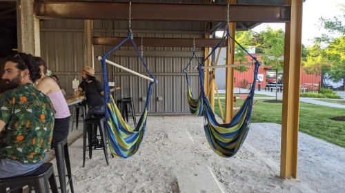 A cozy outdoor space with colorful hammocks hanging, surrounded by people enjoying drinks and a sandy floor.