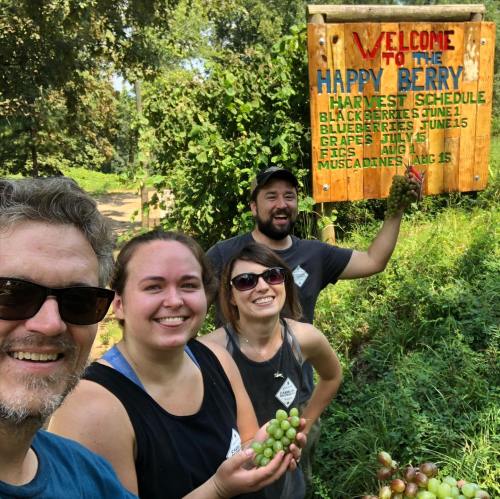 A group of four people smiling in front of a sign for Happy Berry, surrounded by greenery and holding grapes.