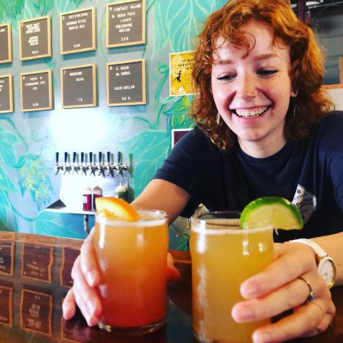 A smiling woman holds two colorful drinks at a bar, with a vibrant wall and drink menu in the background.