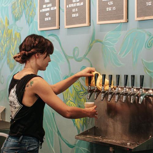 A person pours a drink from a tap at a bar with a colorful wall and menu in the background.