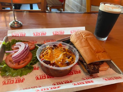 A meal featuring a sandwich, salad with tomato and onion, a side of chili with cheese, and a dark beverage.