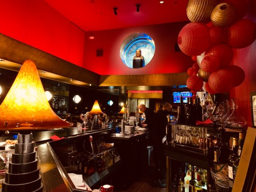 A vibrant bar scene with red lighting, decorative lanterns, and a bartender preparing drinks.