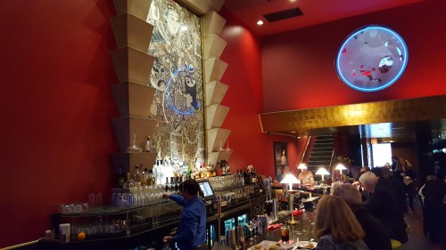 A vibrant bar scene with a red wall, intricate artwork, and patrons enjoying drinks in a stylish setting.