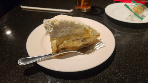 A slice of banana cream pie topped with whipped cream on a plate, with a fork beside it. A drink is in the background.