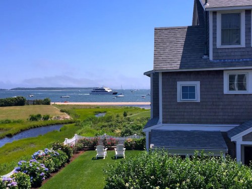 A coastal view featuring a house, green lawn, and boats on the water under a clear blue sky.