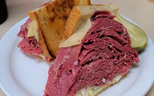 A close-up of a stacked pastrami sandwich on toasted bread, served with pickles on a white plate.