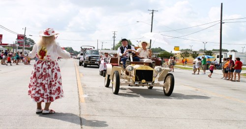A vintage car drives in a parade, with participants in period costumes and a crowd watching under sunny skies.