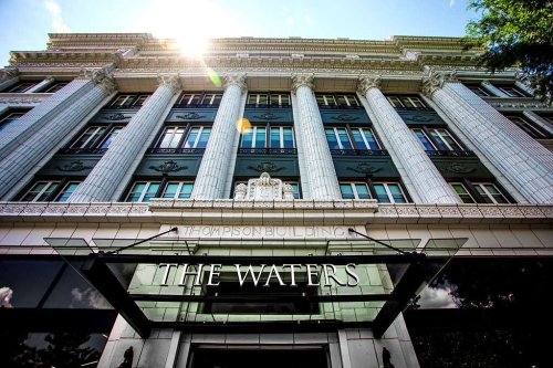 Historic building with tall columns and a glass entrance sign reading "The Waters" under a bright sun.