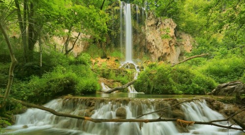 A serene waterfall cascades over rocks, surrounded by lush greenery and a tranquil pool below.