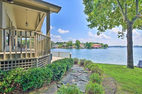 A serene lakeside view featuring a house porch, lush greenery, and calm water under a blue sky with fluffy clouds.