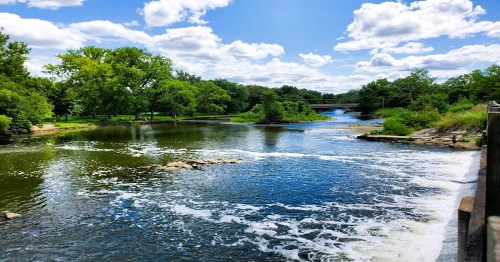 A serene river scene with lush greenery, rocks, and fluffy clouds reflecting in the water under a bright blue sky.