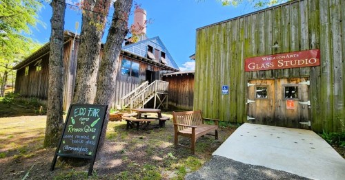 A rustic glass studio with wooden buildings, outdoor seating, and a sign for an eco fair promoting recycled glass.
