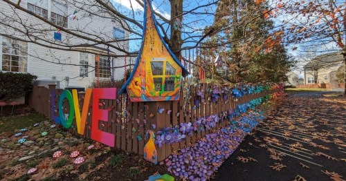 Colorful decorations spell "LOVE" on a wooden fence, surrounded by flowers and vibrant art in a charming outdoor setting.