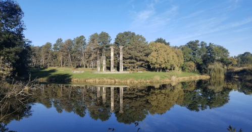 Ruins of tall columns beside a calm lake, surrounded by trees and clear blue skies. Reflections in the water.